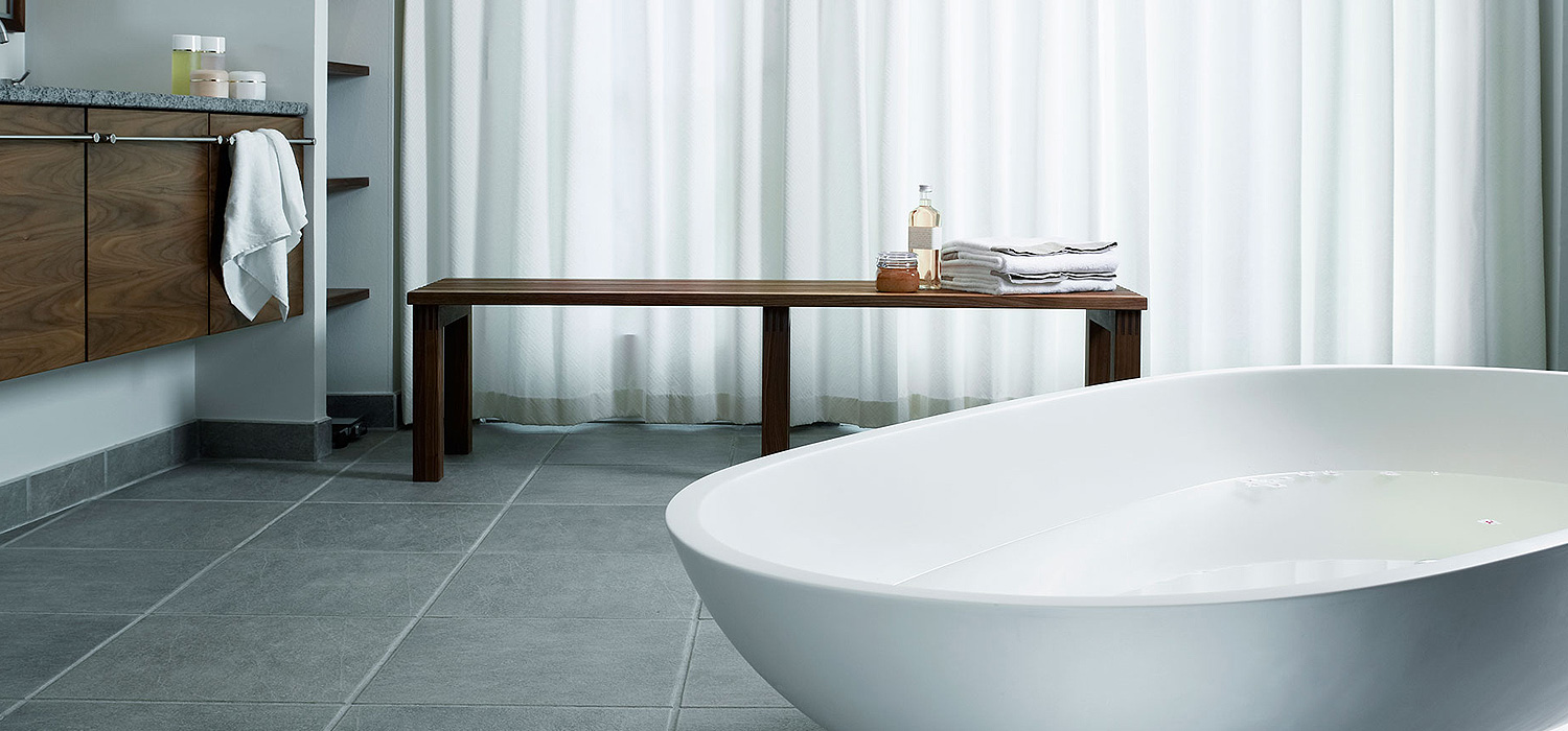 Modernes Badezimmer mit freistehender Badewanne, grauen Bodenfliesen und Holzbank vor lichtdurchlässigem Vorhang.